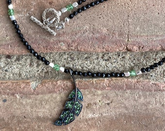 May include: A black beaded necklace with green and silver accents. The necklace features a silver toggle clasp and a pendant in the shape of a leaf, embellished with green stones. The necklace is displayed against a brick background.