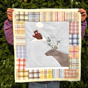 Può includere: Una trapunta fatta a mano con una scena centrale di una renna marrone con corna floreali e una persona con un cappotto marrone su una collina innevata. La trapunta ha un bordo patchwork.
