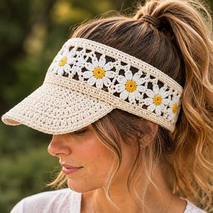 Op de afbeelding: Een beige gehaakte zonneklep met een band van witte madeliefjesmotieven met gele harten. De klep heeft een effen beige rand en een brede, opengewerkte kroonsectie opgebouwd uit onderling verbonden bloempatronen in licht crèmekleurig garen.