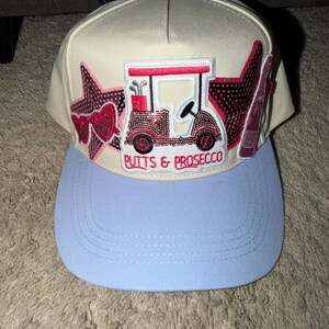 May include: A baseball cap with a light blue brim and a cream-colored crown. The cap features an embroidered golf cart design with the text "Putts & Prosecco". Decorative star and heart accents are also present.