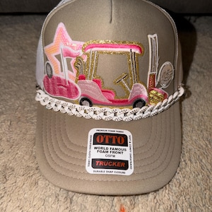 May include: A tan trucker hat with a white mesh back and a golf cart design. The golf cart is pink and gold, with a star and golf clubs. The hat has a white lace trim and a snap closure. The hat has the text "OTTO" and "TRUCKER".