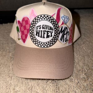 Puede incluir: Gorra de béisbol beige y marrón claro con parches bordados. Los parches incluyen un corazón rosa, un anillo de diamantes y el texto "IT'S GIVING WIFEY" y "MRS". La gorra tiene una visera curva.