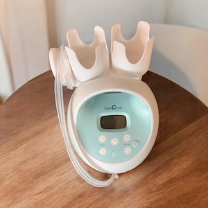 May include: A white and teal Spectra breast pump with a digital display and control buttons. The pump has a unique design with two curved flanges and tubing. The Spectra logo is visible on the front.
