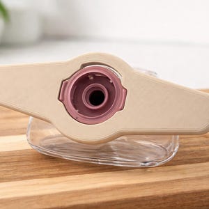 May include: A beige and pink medical device with a clear, glass-like container. The device has a central pink component and a beige, wing-shaped top. It rests on a wooden cutting board.