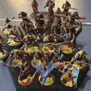 Puede incluir: Figuras en miniatura de guerreros de fantas&iacute;a, pintadas con detalles dorados, azules y rojos. Las figuras est&aacute;n dispuestas en bandejas hexagonales negras, algunas sosteniendo armas y escudos. Las bases est&aacute;n detalladas con hierba y manchas de sangre.
