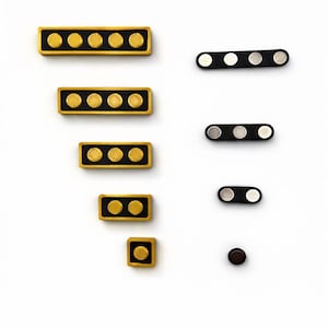 Op de afbeelding: Een verzameling rechthoekige en cirkelvormige magneten in verschillende maten. De rechthoekige magneten hebben een zwarte basis met goudkleurige cirkels, terwijl de cirkelvormige magneten een zwarte basis met zilveren cirkels hebben. Op een witte achtergrond.