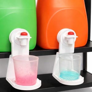 Puede incluir: Contenedores de plástico verde y naranja con dispensadores blancos y botones rojos. Dos vasos de plástico transparente contienen líquido rosa y azul. Los contenedores están en un estante negro.