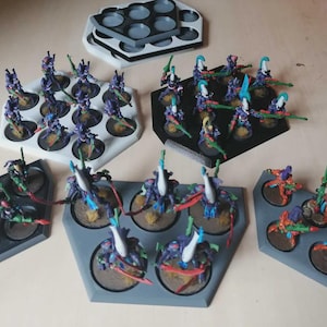 Puede incluir: Figuras en miniatura de varios colores, incluyendo morado, verde y naranja, dispuestas en bandejas hexagonales y circulares. Las figuras est&aacute;n posando con armas y probablemente son para juegos de mesa o coleccionismo. Las bandejas son blancas, negras y grises.