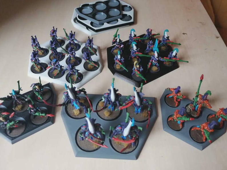 Puede incluir: Figuras en miniatura de varios colores, incluyendo morado, verde y naranja, dispuestas en bandejas hexagonales y circulares. Las figuras est&aacute;n posando con armas y probablemente son para juegos de mesa o coleccionismo. Las bandejas son blancas, negras y grises.
