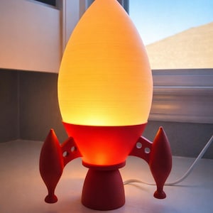 May include: A red and yellow rocket-shaped lamp. The lamp has a red base and fins, with a yellow, glowing, egg-shaped top. The lamp is on a white surface, with a window in the background.