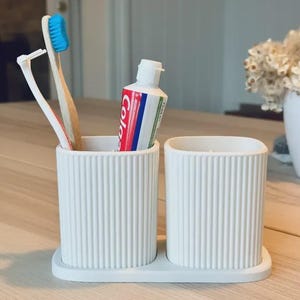 May include: A white ceramic toothbrush holder with a ribbed design, holding a toothbrush, a tongue scraper, and a tube of toothpaste. The holder sits on a matching tray, perfect for bathroom organization.