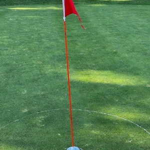 Könnte beinhalten: Eine Golf-Fahne mit roter Flagge. Die Flagge ist an einer langen, orangefarbenen Stange befestigt. Die Stange steckt in einer weißen Kunststoffbasis, die auf einem grünen, grasbewachsenen Feld steht.