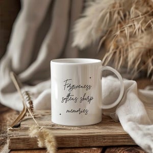 以下が含まれることがあります： 「Forgiveness softens sharp memories」のテキストが黒い筆記体で書かれた白いセラミックマグ。マグカップは湾曲したハンドルを持ち、背景に装飾的な要素のある木製の表面に置かれています。