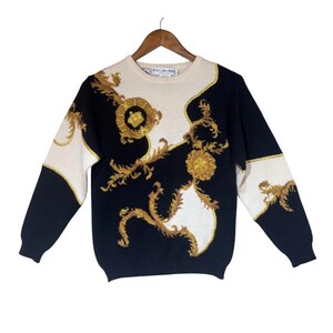 Op de afbeelding: Een vintage trui met een crèmekleurig juk en een gouden barokontwerp op een zwarte achtergrond. De mouwen zijn zwart met crèmekleurige accenten. De trui wordt op een houten hanger gepresenteerd.