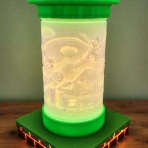 May include: A green and white decorative lamp featuring a detailed relief of a video game scene. The lamp has a cylindrical body with a green top and base, and sits on a green square base with orange accents. The lamp is lit from within.