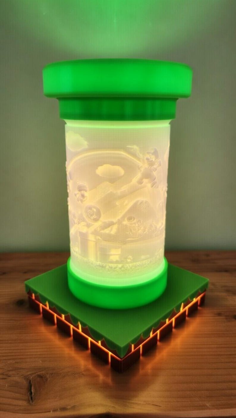 May include: A green and white decorative lamp featuring a detailed relief of a video game scene. The lamp has a cylindrical body with a green top and base, and sits on a green square base with orange accents. The lamp is lit from within.