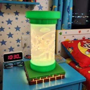 May include: A green and white cylindrical night light with a cartoon scene inside, sitting on a green and brown base. The lamp is on a blue nightstand with a dinosaur sticker. A Pikachu plush and a rocket are on a shelf above.