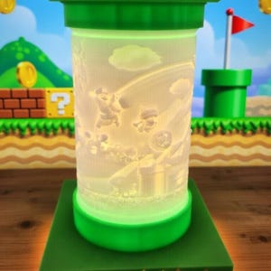 May include: A green and white lamp featuring a Super Mario Bros. theme. The lamp has a cylindrical shape with a green top and base, and a detailed scene of Mario characters. The base has a brick-like design with an orange glow.