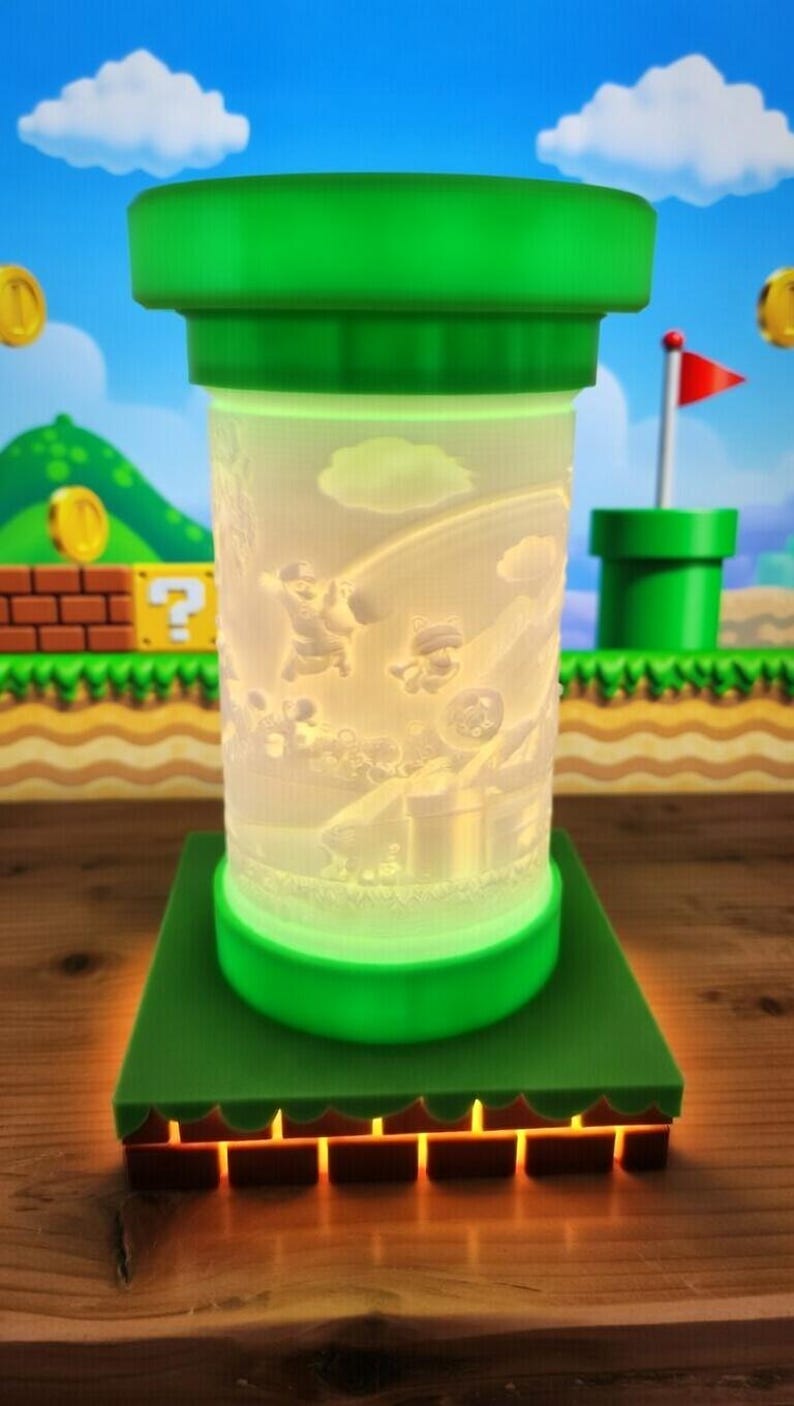 May include: A green and white lamp featuring a Super Mario Bros. theme. The lamp has a cylindrical shape with a green top and base, and a detailed scene of Mario characters. The base has a brick-like design with an orange glow.