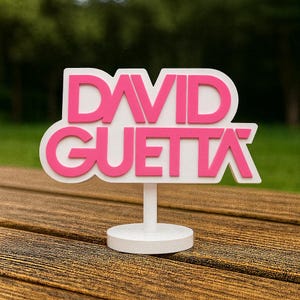 May include: A white and pink sign with the text "DAVID GUETTA" in a bold, stylized font. The sign is mounted on a white stand with a circular base, set against a blurred green background. The sign is likely a decorative item or a piece of merchandise.