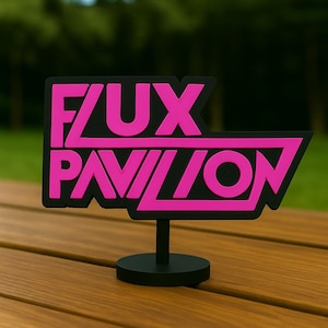 May include: A black sign with the text "FUX PAVILION" in bright pink. The sign is mounted on a small black stand, and is set on a wooden table. The background is blurred green foliage.