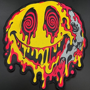 May include: A circular embroidered patch featuring a yellow smiley face with red spiral eyes and a wide, toothy grin. The design includes red and yellow liquid-like drips running down the face, with a grey skull integrated into the right side.