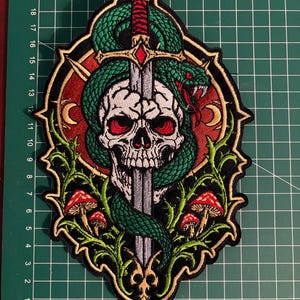 May include: An embroidered patch featuring a white skull pierced by a silver sword, wrapped in a green snake with red eyes. The design includes red mushrooms, green vines, and gold crescent moons on a red background, measuring 22 cm in height.