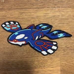 Peut inclure: Écusson brodé représentant une créature aquatique bleue avec des détails blancs, rouges et bleu clair. Le patch a une apparence détaillée et stylisée, avec une bordure noire. Le motif est posé sur un fond en bois.