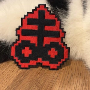 Peut inclure: Un écusson pixelisé rouge et noir représentant une croix stylisée. L'écusson est posé sur une surface en bois, avec un arrière-plan flou de fourrure noire et blanche. L'écusson a une esthétique rétro et numérique.