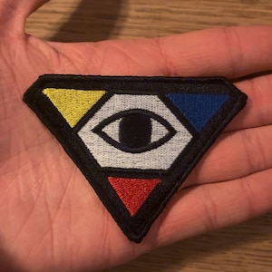 May include: A black, diamond-shaped embroidered patch featuring a stylized eye design. The eye is surrounded by yellow, blue, and red triangles. The patch is held in a hand.