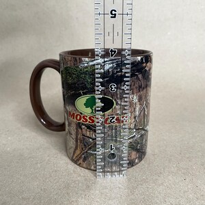 May include: A brown ceramic mug with a camouflage pattern and a brown handle. The mug features the "Mossy Oak" logo in green and white. The mug is approximately 4 inches tall.