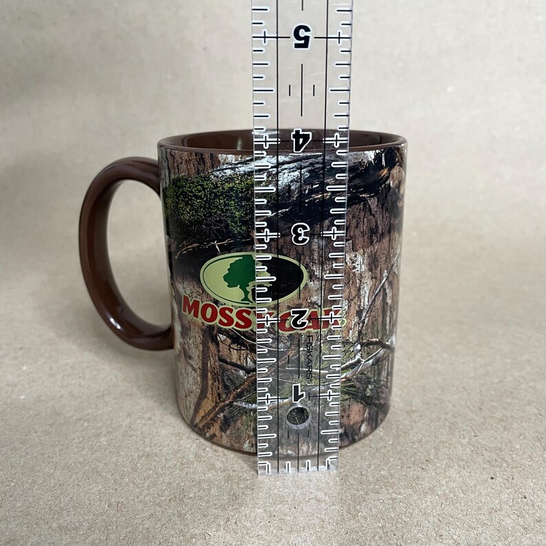 May include: A brown ceramic mug with a camouflage pattern and a brown handle. The mug features the "Mossy Oak" logo in green and white. The mug is approximately 4 inches tall.