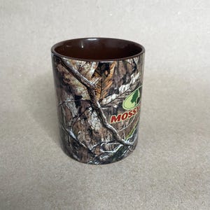 May include: A cylindrical tumbler with a brown interior and a camouflage exterior featuring a tree bark pattern. The tumbler has the word "Mossy" in a green and white logo.