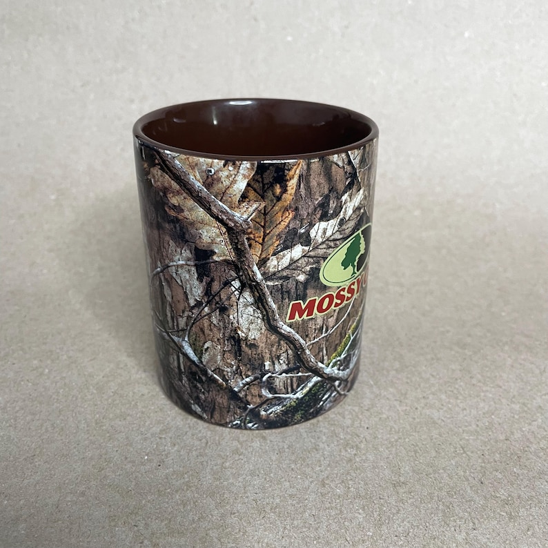 May include: A cylindrical tumbler with a brown interior and a camouflage exterior featuring a tree bark pattern. The tumbler has the word "Mossy" in a green and white logo.
