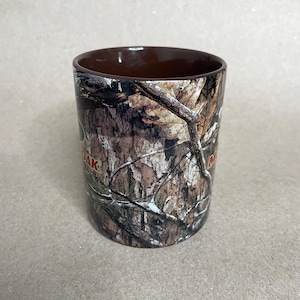 May include: A cylindrical ceramic mug with a brown interior and a camouflage pattern on the exterior. The camouflage design features shades of brown, tan, and green, with the word "Mossy Oak" in red.