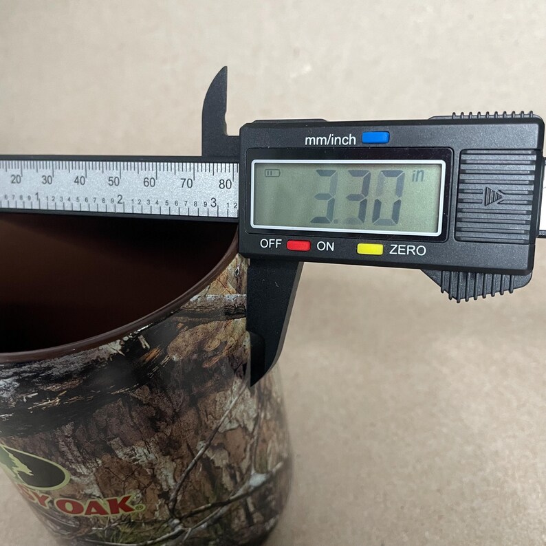 May include: A digital caliper measures the diameter of a brown camouflage-patterned tumbler. The digital display reads 3.30 inches. The tumbler has the Mossy Oak logo.