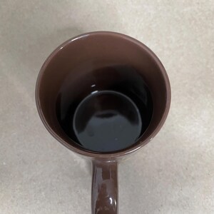 May include: A brown ceramic mug with a handle, viewed from above. The interior of the mug is dark, possibly filled with a liquid. The mug is a solid, uniform color.
