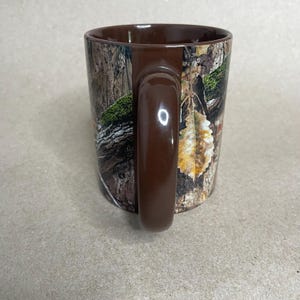 May include: A brown ceramic mug with a camouflage pattern. The mug features a dark brown handle and rim. The camouflage design includes shades of brown, green, and tan, resembling a forest scene. The mug is designed for hot or cold beverages.