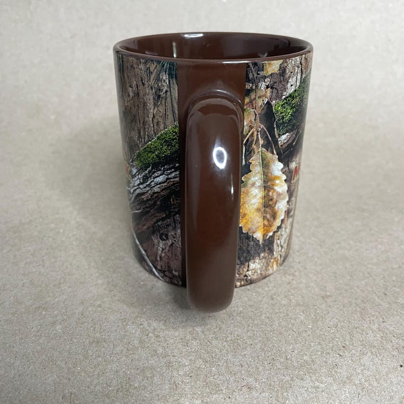 May include: A brown ceramic mug with a camouflage pattern. The mug features a dark brown handle and rim. The camouflage design includes shades of brown, green, and tan, resembling a forest scene. The mug is designed for hot or cold beverages.