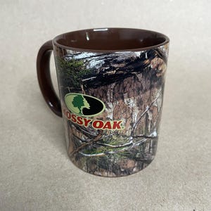 May include: A ceramic coffee mug with a brown interior and handle. The exterior features a camouflage pattern with the "Mossy Oak" logo in green, black, and white. The mug is designed for hot beverages.
