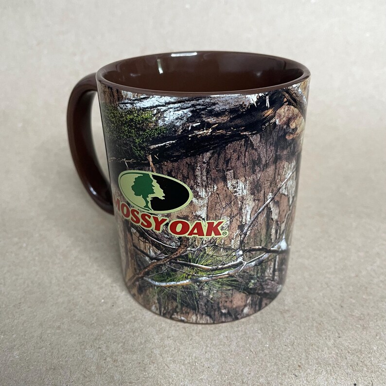 May include: A ceramic coffee mug with a brown interior and handle. The exterior features a camouflage pattern with the "Mossy Oak" logo in green, black, and white. The mug is designed for hot beverages.