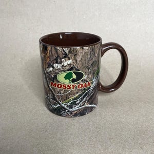 May include: A brown ceramic mug with a camouflage pattern and a brown handle. The mug features the "Mossy Oak" logo in green, white, and red. The mug is designed for hot or cold beverages.