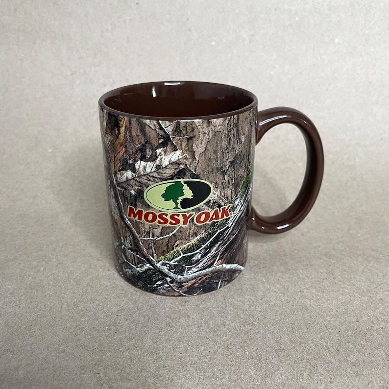 May include: A brown ceramic mug with a camouflage pattern and a brown handle. The mug features the "Mossy Oak" logo in green, white, and red. The mug is designed for hot or cold beverages.