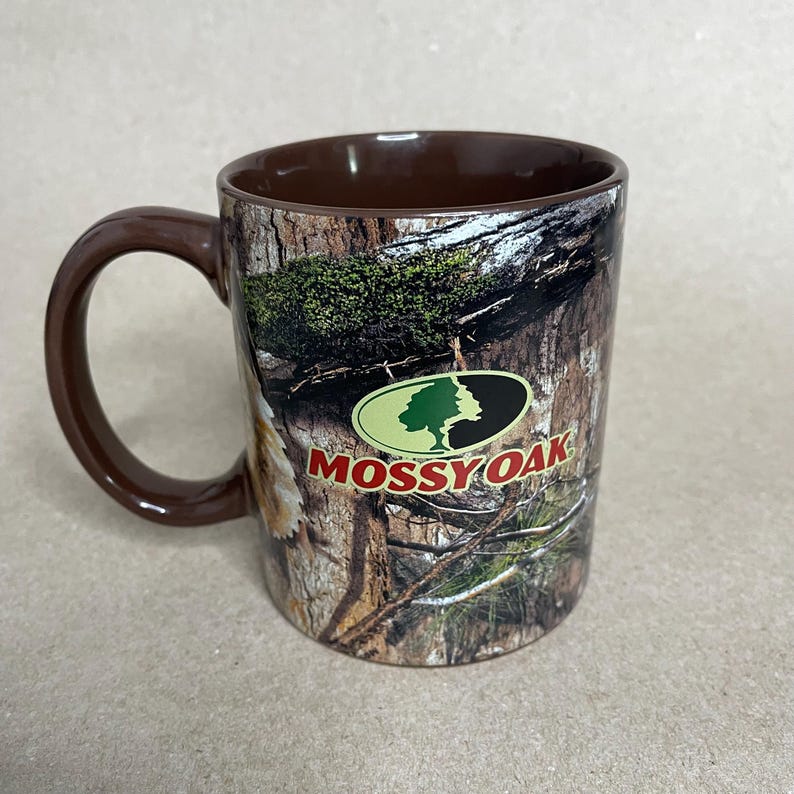 May include: A brown ceramic mug with a camouflage pattern and a brown handle. The mug features the "Mossy Oak" logo in green and red. The interior of the mug is also brown.