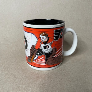 May include: White ceramic mug featuring a hockey goalie in orange and black uniform. The mug has a black interior and a white handle. The design includes the team logo and a hockey rink graphic.