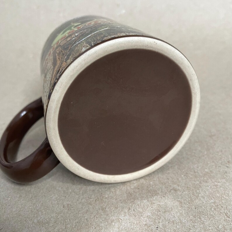 May include: A brown ceramic mug with a camouflage pattern. The mug has a dark brown handle and a light beige rim. The bottom of the mug is a solid dark brown color.