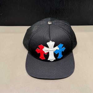 May include: A black trucker hat featuring a solid black front panel and a mesh back. The front displays three embroidered cross patches in red, white, and blue. A small silver-toned metallic button is centered at the top of the crown.