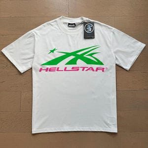 Könnte beinhalten: Weißes T-Shirt mit einem grün-rosa Grafikdesign. Das Design zeigt einen Stern und abgewinkelte Linien, mit dem Wort "HELLSTAR" in Pink. Ein schwarzes Etikett mit Logo ist am Kragen befestigt.