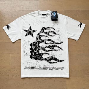 Könnte beinhalten: Weißes T-Shirt mit einem schwarz-weißen Grafikdesign. Das Design zeigt einen Stern, eine flammenartige Silhouette und das Wort "HELLSTAR". Das T-Shirt hat ein angebrachtes Etikett.