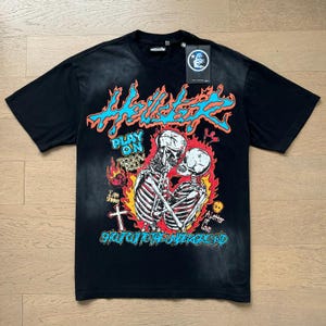 Pode incluir: Camiseta preta com um gráfico de dois esqueletos abraçados, rodeados por chamas. A camiseta apresenta o texto "Hellstar" em azul e laranja, juntamente com outras frases como "Play On" e "Shot Out To The Underground."
